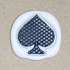 May include: A white wax seal featuring a black spade symbol. The spade design has a textured pattern. The seal is set against a light brown background.