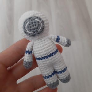 May include: A small, handmade crochet astronaut doll. The astronaut is primarily white with gray accents on the helmet, hands, and feet. Blue stripes adorn the arms and legs. The doll is held in a hand.