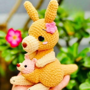May include: A crocheted kangaroo toy in tan and cream, with a pink flower. The kangaroo holds a smaller pink joey. The toy has black eyes with green highlights, set against a blurred green background. A handmade soft toy.