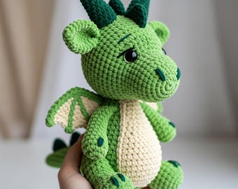 May include: A crocheted dragon toy in green and cream. The dragon has large eyes, small horns, and wings. The soft toy is ideal for a child's room or as a unique gift. The dragon is made with soft yarn.