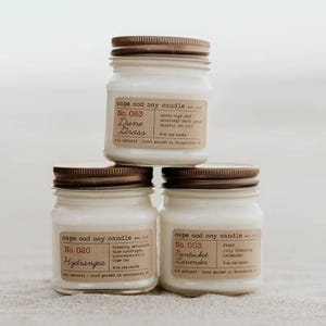 May include: Three glass jars with brown lids and labels. The labels are for Cape Cod soy candles. The scents are Dune Grass, Hydrangea, and Nantucket Lavender. Each jar is 9 oz.