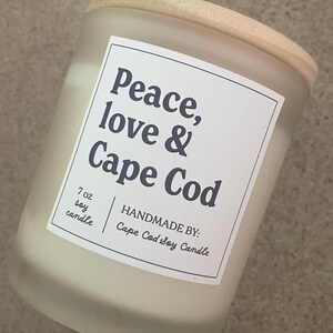 May include: A 7 oz soy candle in a frosted glass jar with a light wood lid. The white rectangular label features black text that reads: Peace, love & Cape Cod, 7 oz soy candle, and HANDMADE BY: Cape Cod Soy Candle, enclosed in a thin black border.