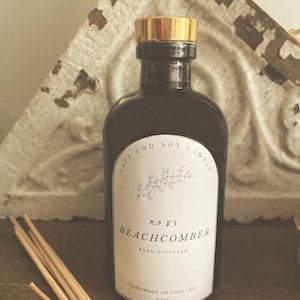 May include: A dark glass bottle with a gold cap, labeled "Cape Cod Soy Candle" and "Beachcomber Reed Diffuser." The label includes botanical illustrations and the text "Handmade on Cape Cod" and "9 oz.". Reed sticks are in the foreground.