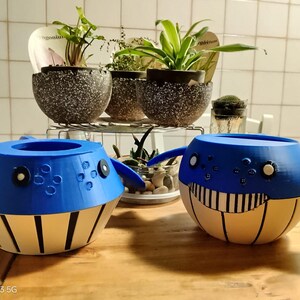 May include: Two blue and white whale-shaped planters with black accents are on a wooden surface. The planters have a round body with a wide opening and fin-like extensions. Behind them are several potted plants on a metal rack.