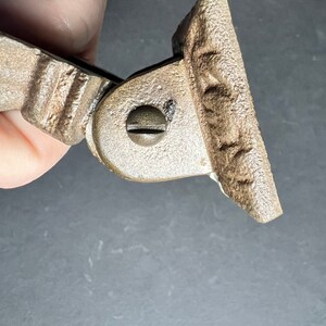 May include: A close-up of a metal hinge with a decorative design. The hinge is made of a light brown metal and has a screw in the center.