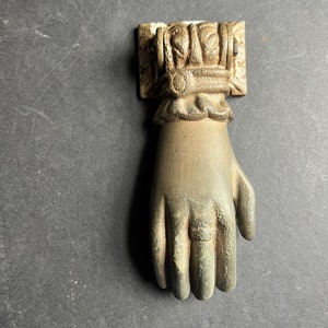 May include: A gold-toned metal door knocker shaped like a hand with a detailed design. The knocker is mounted on a square base with decorative accents.