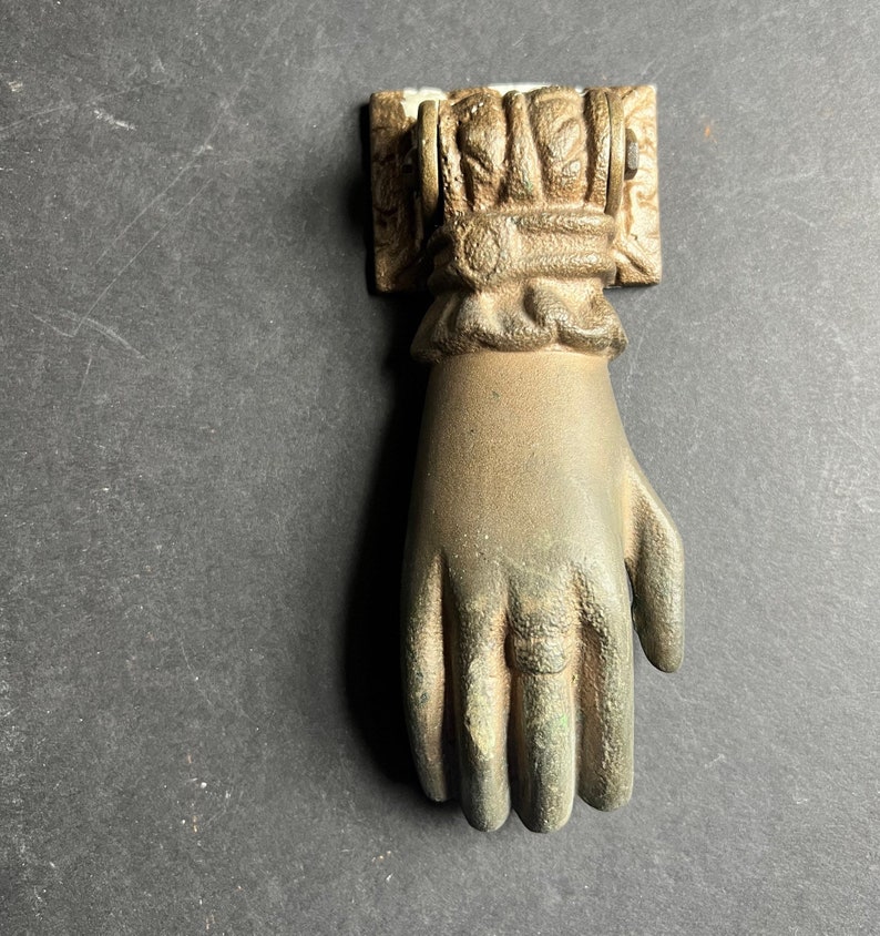 May include: A gold-toned metal door knocker shaped like a hand with a detailed design. The knocker is mounted on a square base with decorative accents.