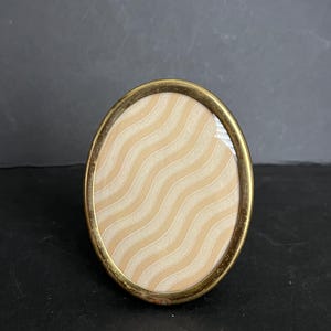 May include: An oval-shaped picture frame with a metallic gold-toned finish. The frame contains a decorative insert featuring a repeating pattern of light beige and cream-colored vertical wavy lines that span the entire surface of the oval interior.