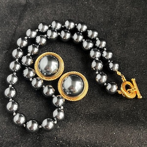 May include: A dark grey beaded necklace and matching earrings set. The necklace features large, round beads and a gold-tone clasp. The earrings have a gold-tone frame around a large, round, dark grey bead.