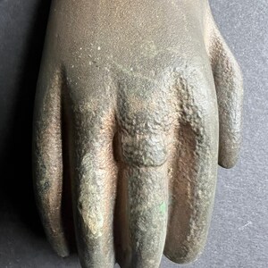 May include: A close-up of a cast iron hand door knocker. The knocker is a realistic depiction of a hand with a ring on the index finger. The hand is a dark brown color with a green patina.
