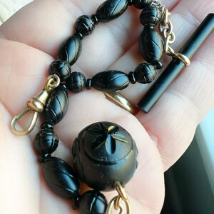 May include: A black jet black onyx watch chain with a gold clasp and a large, round, carved onyx fob. The chain is made of oval-shaped beads.