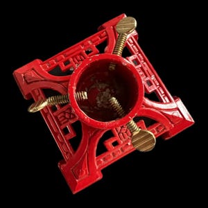 May include: A red metal Christmas tree stand with a square base and a central cylindrical holder. The stand features decorative cutouts and gold-colored screw fasteners for securing the tree. The stand is designed to hold a Christmas tree.