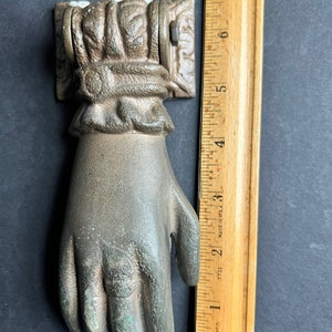 May include: A bronze hand-shaped door knocker with intricate details. The knocker is attached to a square plate with ornate designs. The knocker is approximately 6 inches tall.