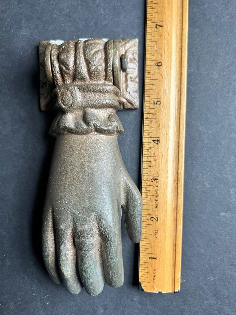 May include: A bronze hand-shaped door knocker with intricate details. The knocker is attached to a square plate with ornate designs. The knocker is approximately 6 inches tall.