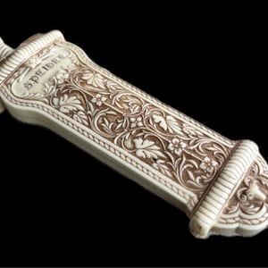 May include: Ivory-colored decorative object with intricate floral carvings and the word "SPEIDER" etched on the surface. The object has a rectangular shape with rounded ends and a detailed, textured design.