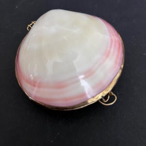 May include: A polished clam shell locket featuring a smooth, cream-colored center with soft pink concentric bands. The shell is encased in a thin gold-toned metal frame with a small hinge on one side and a circular jump ring attached to the edge.