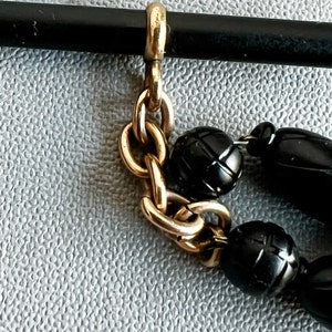 May include: A gold chain necklace with black beads. The chain is made of small, linked circles. The beads are round and have a slightly textured surface.