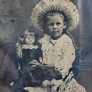 Könnte beinhalten: Altes Schwarz-Weiß-Foto eines jungen Mädchens mit großem, verziertem Hut, das eine Puppe hält. Die Puppe trägt ein dunkles Kleid mit weißem Kragen. Das Foto hat ein gealtertes, abgenutztes Aussehen.