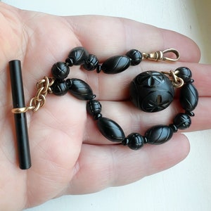 May include: A black onyx watch chain with a gold chain and a carved black onyx fob. The chain is made of round beads and the fob is a large, round bead with a carved design.