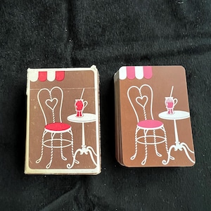 May include: Two decks of playing cards with a brown cafe design. Each card features a heart-shaped chair, a small table with a drink, and a striped awning. The cards are stacked on a black surface.
