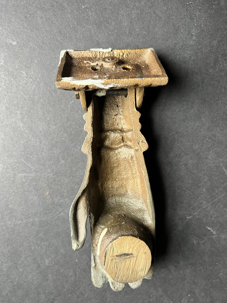 May include: A vintage brass door knocker shaped like a hand. The knocker has a wooden base and a metal hand with a hinged arm.