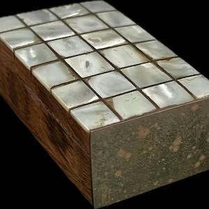 Può includere: Una scatola rettangolare in legno con coperchio intarsiato in madreperla. Il coperchio è composto da piastrelle quadrate e la scatola ha un bordo metallico. Il legno è di un ricco colore marrone scuro e le piastrelle di madreperla sono iridescenti.