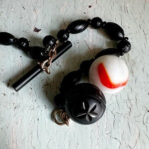 May include: A black beaded necklace with a toggle clasp and a large white and red glass bead. The necklace is made of black beads with a carved design.