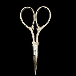 May include: A pair of silver-toned metal embroidery scissors featuring ornate, embossed scrollwork patterns on the handles and finger loops. The blades are pointed and straight, with small, circular hallmark stamps visible near the pivot point.