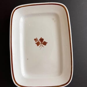 May include: A rectangular white ceramic serving dish with rounded corners. The dish features a thin brown painted border along the rim and a small, detailed brown botanical leaf illustration centered on the interior base of the vessel.
