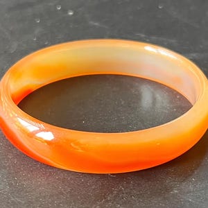 May include: A smooth, polished bangle bracelet crafted from translucent orange carnelian stone. The circular jewelry piece features natural color variations ranging from deep reddish-orange to lighter, pale yellow-orange tones throughout the band.