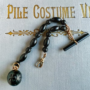May include: A black onyx watch chain with a gold clasp and a black onyx fob. The chain is made of round beads with a carved design.