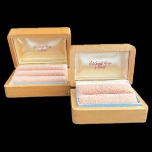 Puede incluir: Dos joyeros de madera abiertos con soportes de anillos de terciopelo rosa claro. Las cajas tienen un exterior de color beige claro y un forro interior color crema. El texto "Art-Carved Rings by Wood" está impreso en rojo en el interior de la tapa.