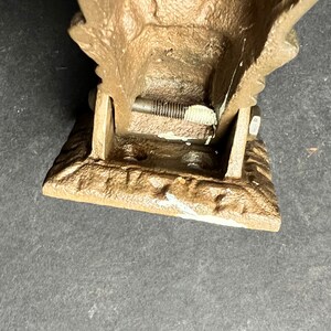May include: A cast iron doorstop in the shape of a stylized eagle with a brown patina. The doorstop is hinged and has a metal screw visible in the hinge.