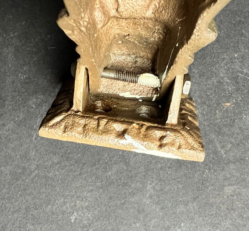 May include: A cast iron doorstop in the shape of a stylized eagle with a brown patina. The doorstop is hinged and has a metal screw visible in the hinge.