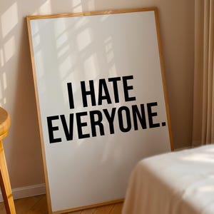 May include: A framed white poster with the text "I HATE EVERYONE" in bold black letters. The poster is leaning against a wall, with a wooden stool and a bed visible in the background. The frame is made of light-coloured wood.