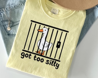 May include: A pale yellow t-shirt featuring a graphic of a white duck with an orange beak and feet inside a black cage. Below the cage, the text reads "got too silly" in a bold, rounded black font. The shirt is displayed on a flat surface.