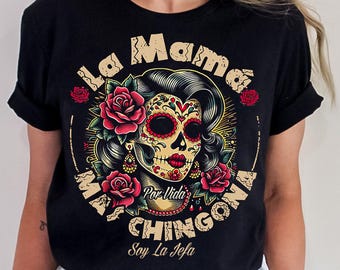 Può includere: Una t-shirt nera con la grafica di una donna teschio di zucchero con rose rosse, orecchini dorati e una collana. Il testo recita La Mama Mas Chingona, Por Vida e Soy La Jefa. Il design è circolare con uno sfondo a raggi di sole gialli.