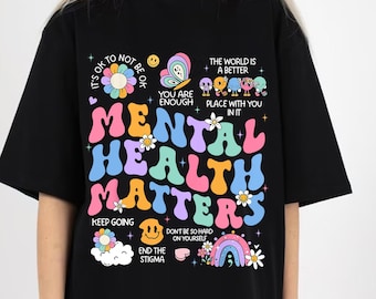 May include: Black t-shirt with a colorful graphic that says "Mental Health Matters". The graphic includes flowers, butterflies, and smiley faces. The text "It's OK to not be OK", "You are enough", "The world is a better place with you in it", "Keep going", "Don't be so hard on yourself", and "End the stigma" is also included in the graphic.