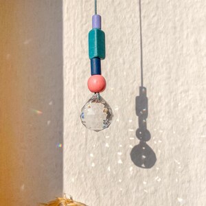 Könnte beinhalten: Ein Kristall-Sonnenfänger mit einem klaren Glasprisma, das an einer Schnur mit bunten Perlen hängt. Die Perlen sind lila, türkis, dunkelblau und rosa.