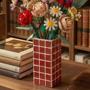 May include: A colourful flower arrangement in a red, square-patterned vase. The bouquet features red roses, pink peonies, and white daisies. The vase is on a wooden desk in front of a bookshelf filled with books.