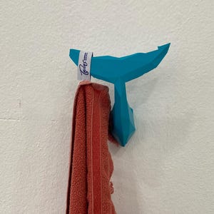 May include: A teal whale tail-shaped wall hook with a coral-coloured towel hanging from it. The hook is mounted on a white textured wall. The towel has a white tag with the word "Taylor's" on it.