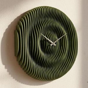 Pode incluir: Um relógio de parede moderno, verde-oliva, com um design em camadas único. O relógio apresenta um mostrador circular com um padrão ondulado e ponteiros prateados. Está montado numa parede branca. Uma pequena suculenta, livros e uma chávena de café estão numa mesa de madeira.