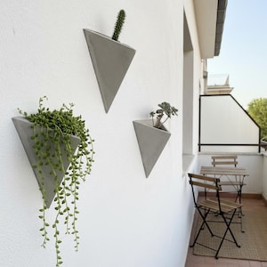 Może przedstawiać: Trzy szare, trójkątne donice ścienne z różnymi zielonymi roślinami. Jedna zawiera kaktus, druga roślinę zwisającą, a trzecia małą sukulent. Donice są zamontowane na białej ścianie, tworząc nowoczesną, minimalistyczną estetykę.