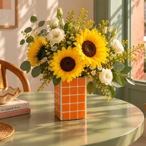 May include: A square ceramic vase with a bright orange grid pattern holds a vibrant floral arrangement featuring large yellow sunflowers, white lisianthus, small yellow filler flowers, and green eucalyptus leaves, creating a cheerful display.