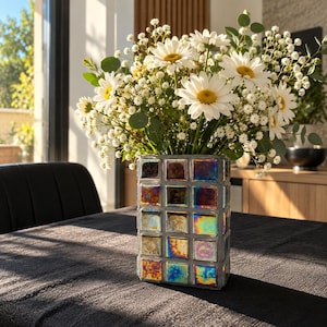 May include: A rectangular vase featuring a grid of iridescent, multi-colored square tiles with a metallic finish, filled with a large bouquet of white daisies and small white baby's breath flowers with green eucalyptus leaves.