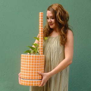 May include: A cylindrical orange planter featuring a white grid pattern and a matching vertical moss pole extension. The planter contains a green leafy plant with pointed foliage. The design consists of small square tiles arranged in a uniform grid.