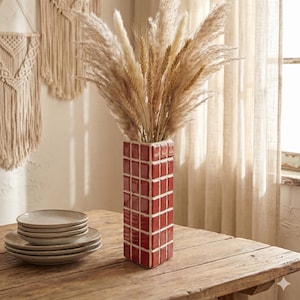 May include: A red tiled vase, filled with dried pampas grass and wheat, stands on a wooden table. Beside it are stacked beige plates. The vase's rectangular shape is a striking contrast to the soft, natural textures of the dried flowers. A macrame wall hanging is in the background.