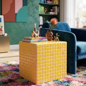 May include: A square side table covered in small, glossy yellow tiles. The table surface holds two stacked books with the text ARTIYO visible on the spine, a tall amber-colored glass vase containing a single orange flower, and a small gold figurine.