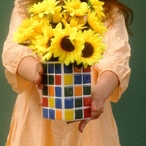 May include: A square vase decorated with a grid of small, colourful mosaic tiles in shades of red, blue, yellow, orange, and green. The vase is filled with a bouquet of bright yellow artificial sunflowers with dark brown centres.