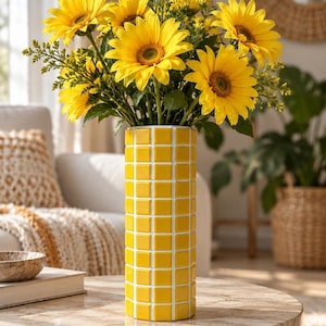 May include: A cylindrical vase featuring a bright yellow grid pattern with white grout lines, filled with a large bouquet of vibrant yellow sunflowers and small green filler flowers, standing upright on a smooth, circular stone-topped table.